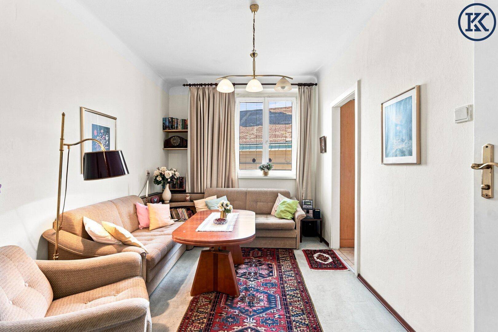 Wohnzimmer mit beigem Sofa, Holztisch und einem bunten Teppich auf hellem Boden.
