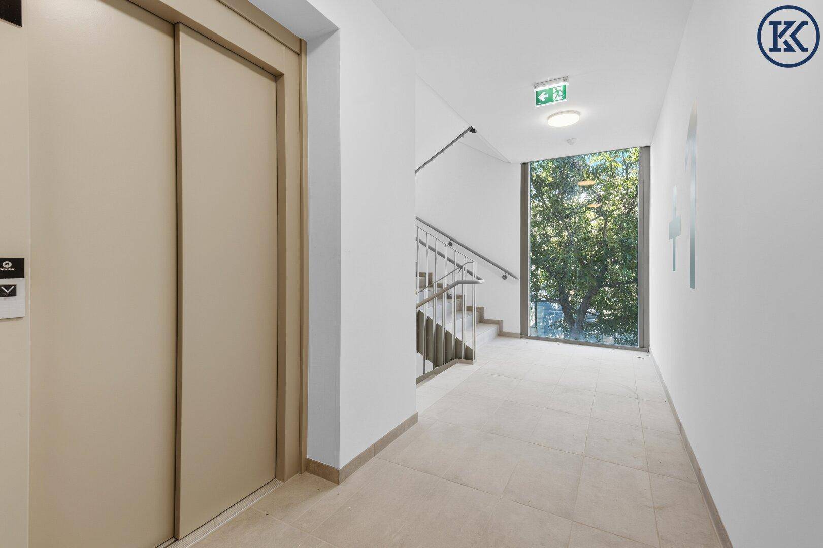 Moderner Flur mit Aufzug, Treppenhaus und großem Fenster mit Blick ins Grüne.