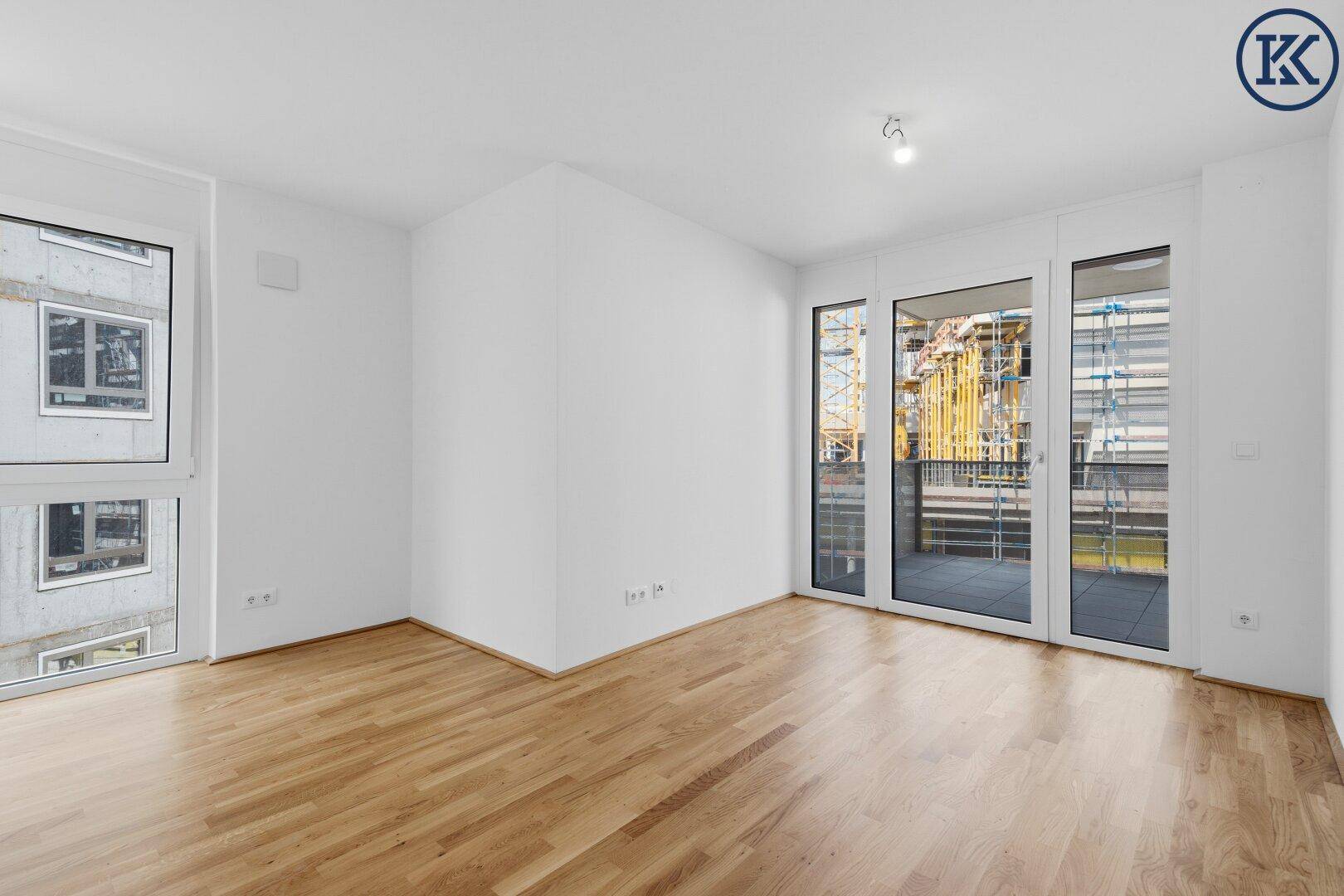 Geräumiger, leerer Raum mit hellem Parkettboden und großen Fenstern zum Balkon.
