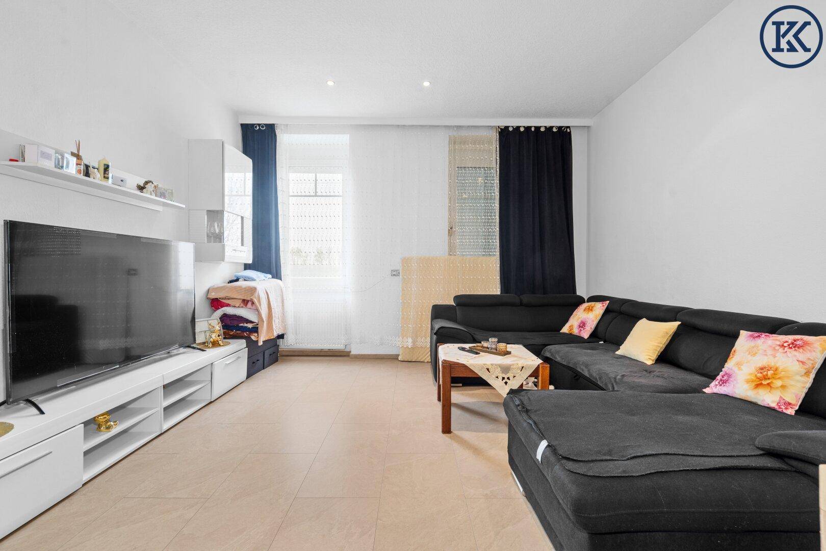 Helles Wohnzimmer mit großem Fernseher, Ecksofa und Fenstern mit dunklen Vorhängen.