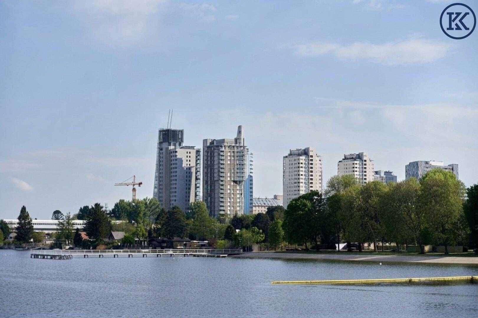 Weitläufiger Blick über den See auf die städtische Skyline mit modernen Gebäuden und einem Baukran am Horizont.