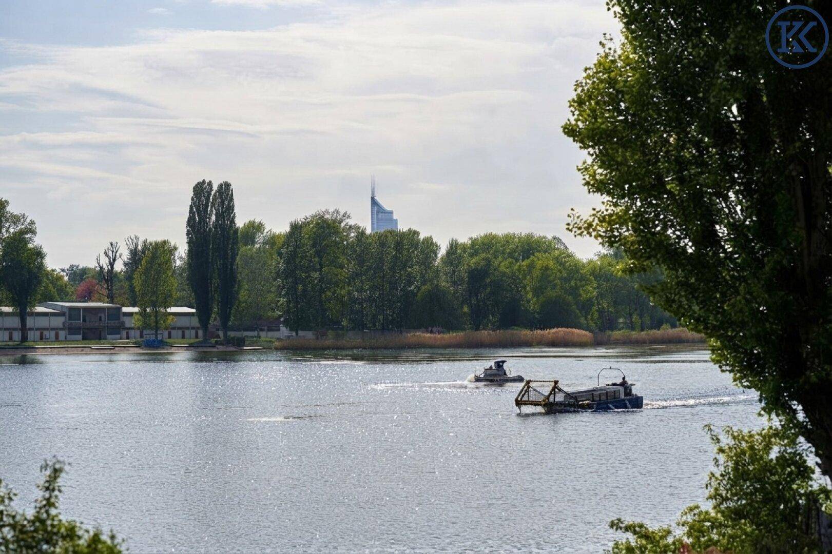 Ruhiger See mit zwei Booten und grünen Ufern, im Hintergrund ist ein markantes Hochhaus der Stadt zu sehen.
