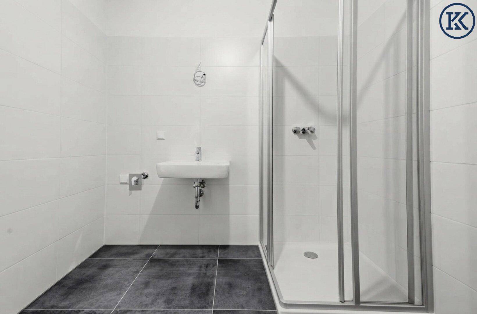 Helles Badezimmer mit moderner Duschkabine und Waschbecken, kombiniert mit dunklen Bodenfliesen.