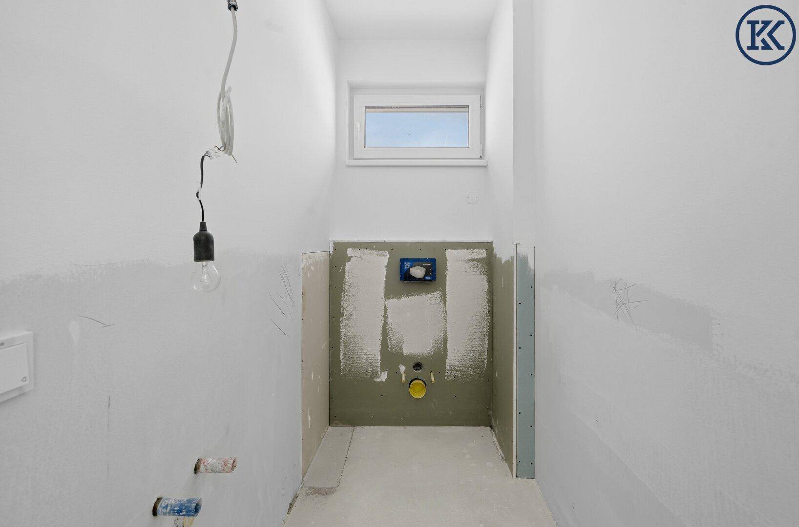 Kleiner Raum im Rohbau mit Fenster und vorbereiteten Anschlüssen für ein WC oder Badezimmer.