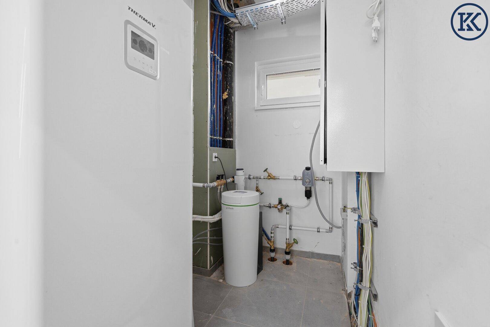 Technikraum mit moderner Wärmepumpe, Leitungen und kleinem Fenster.