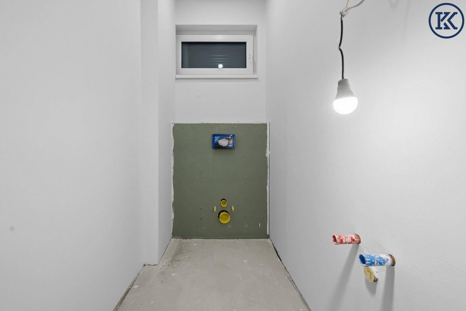Unfertiger WC-Raum mit Fenster und vorbereiteten Anschlüssen für Toilette und Waschbecken.