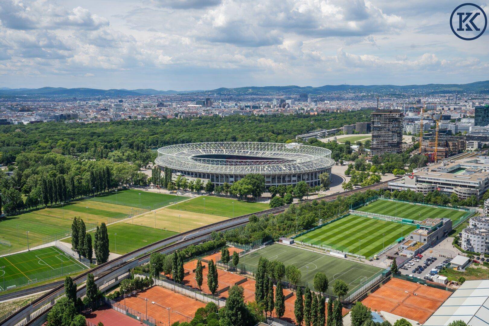 Weitläufiger Blick über die Stadt mit einem großen Stadion und Sportfeldern unter blauem Himmel.