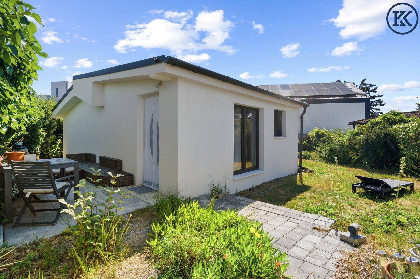 Seitenansicht des Hauses mit einer gepflasterten Terrasse, Gartenmöbeln und einem grünen Garten.