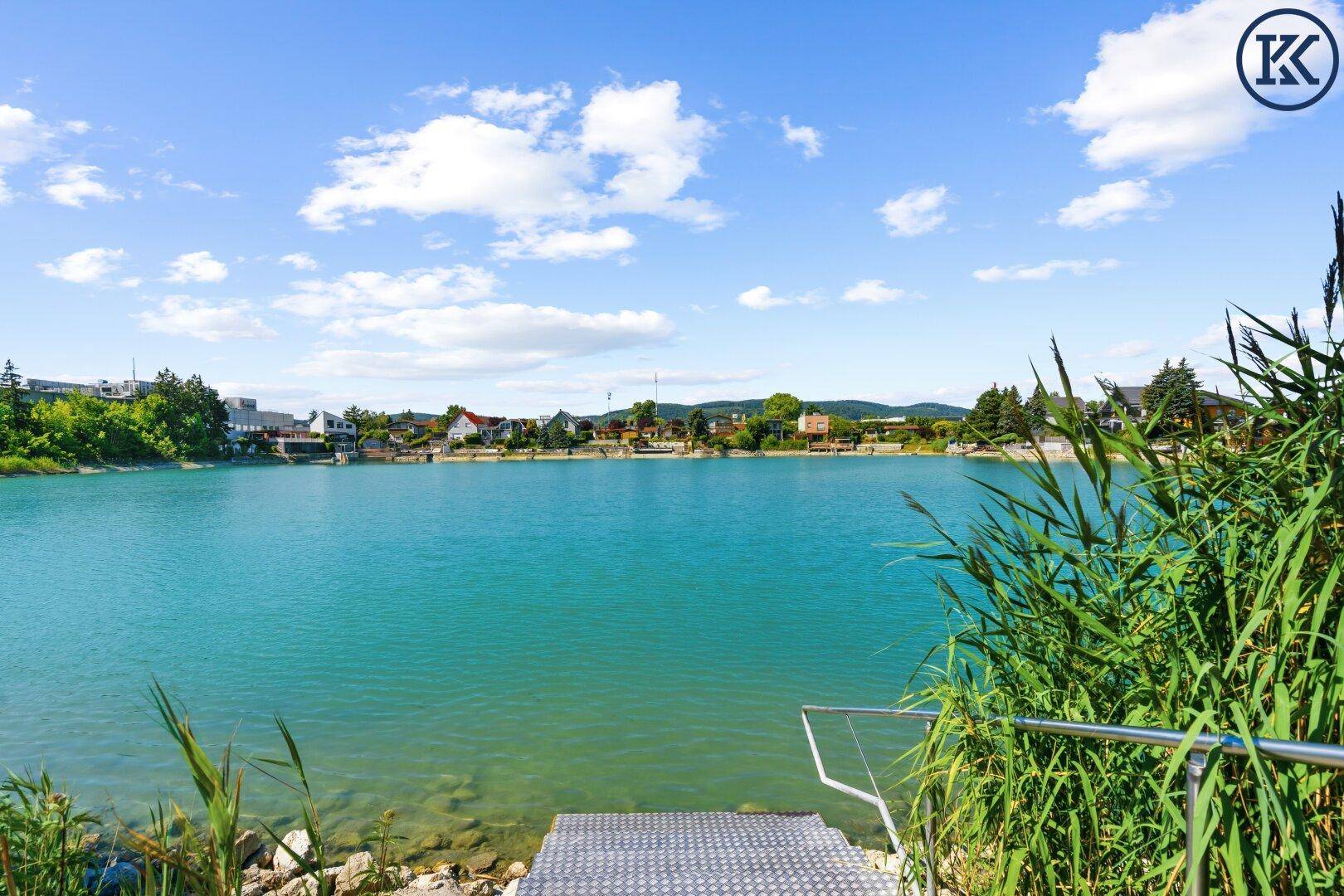 Blick auf einen ruhigen See mit Schilf im Vordergrund und einer kleinen Badeplattform.