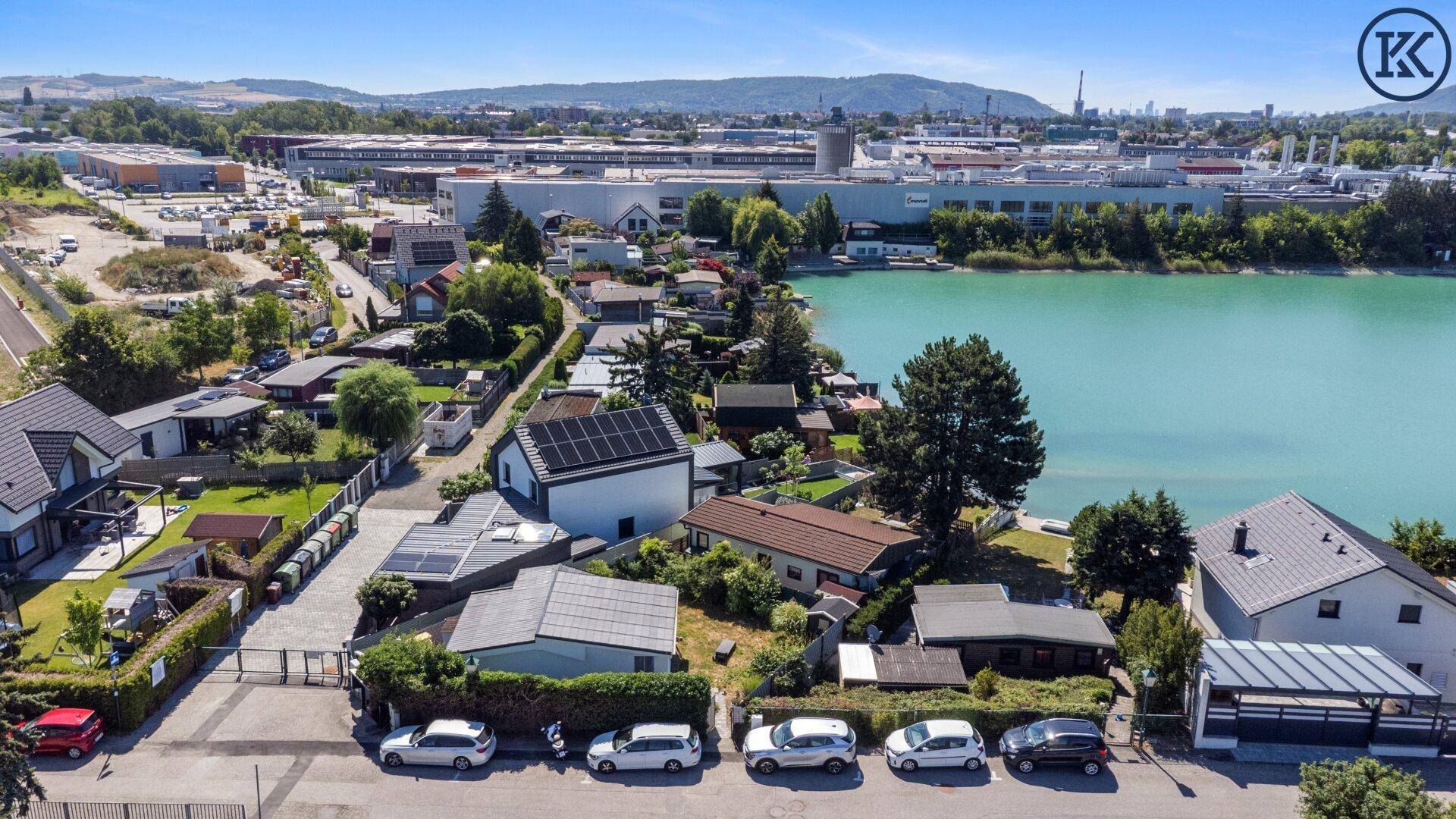 Luftaufnahme von Wohnhäusern und Gärten am Seeufer mit Industriegebäuden in der Ferne.