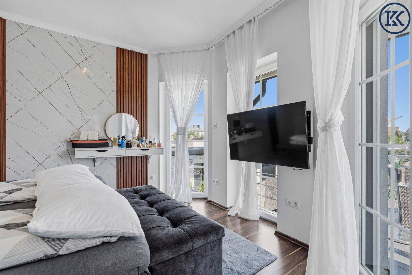 Helles Schlafzimmer mit Wandfernseher, großen Fenstern und leichten Vorhängen für viel Licht.