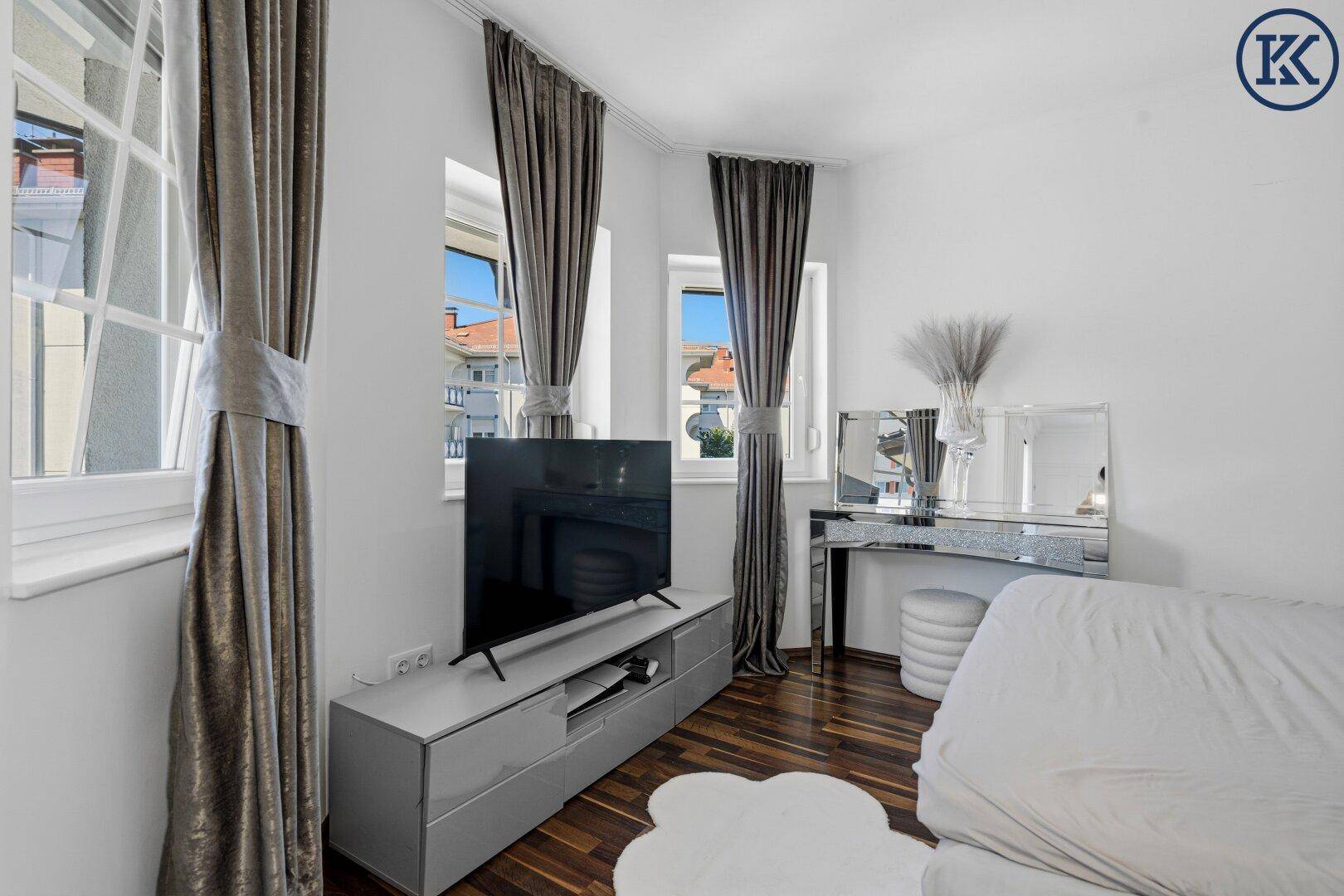 Helles Schlafzimmer mit Fernseher, dunklen Vorhängen und Blick nach draußen.