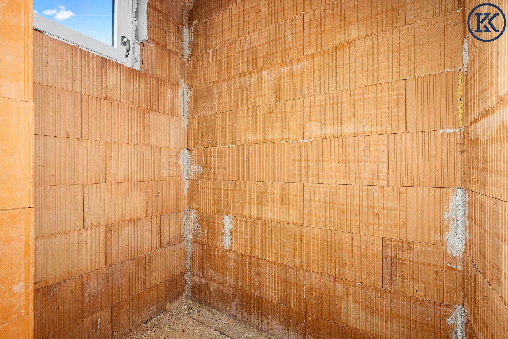 Kleiner Raum im Rohbau mit Ziegelwänden und einem Fenster, das Tageslicht hereinlässt.