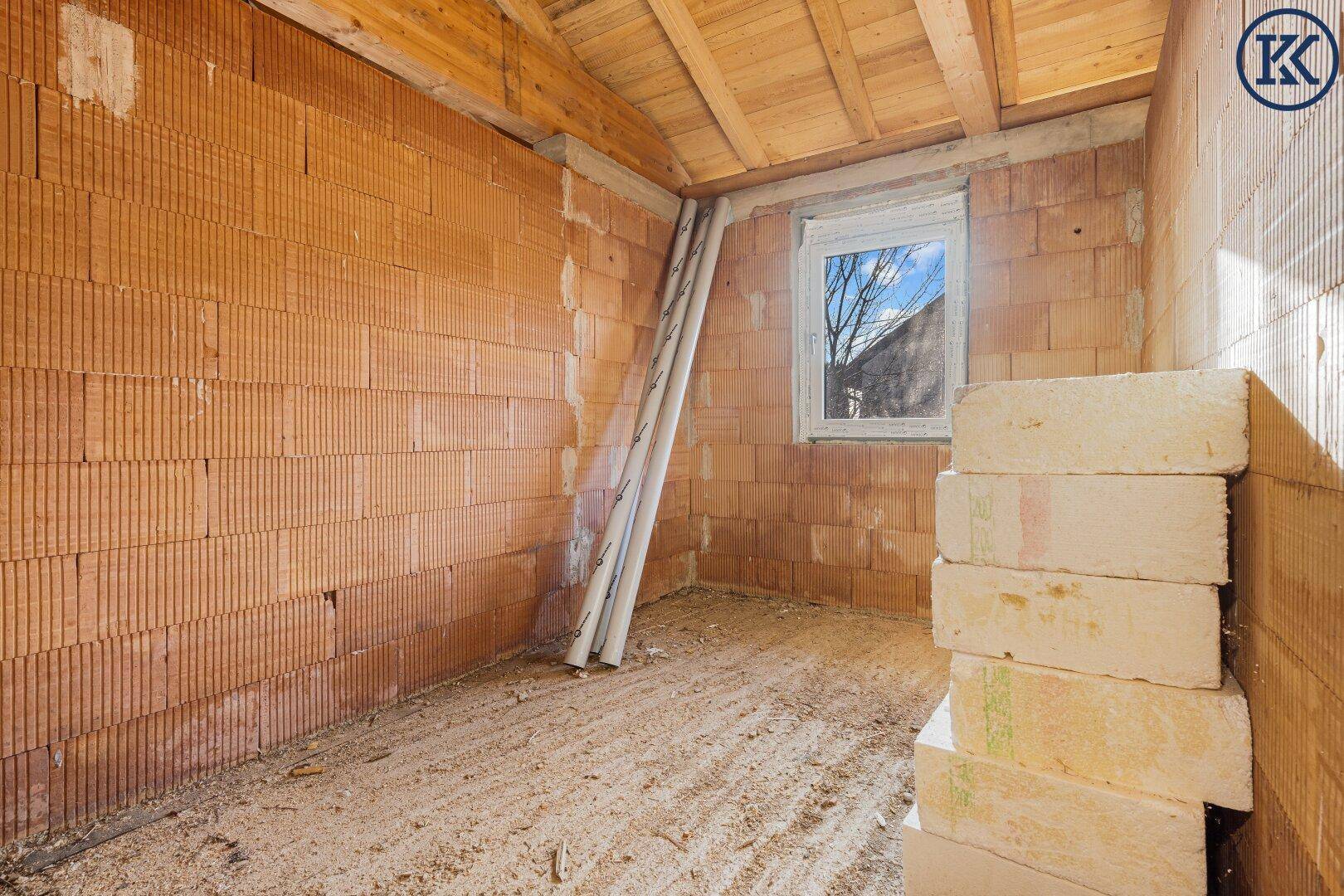 Rohbau eines Zimmers mit Fenster und gestapelten Baumaterialien unter der Holzdecke.