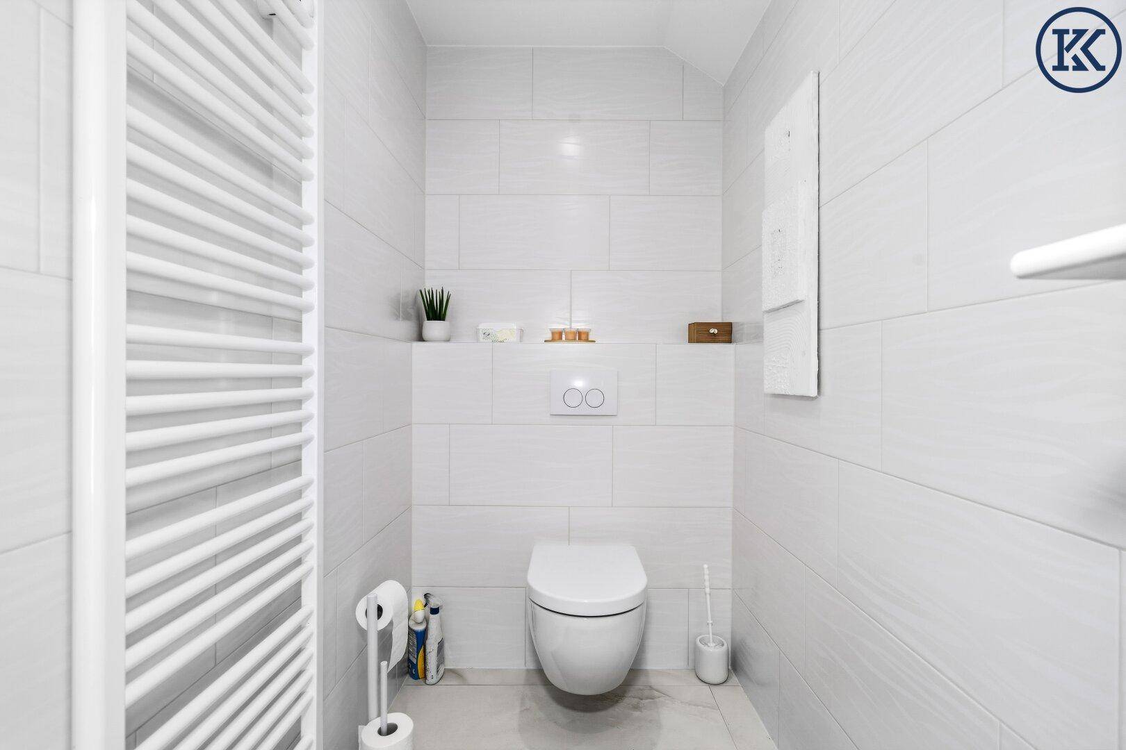 Separates WC mit wandhängender Toilette, weißen Fliesen und Handtuchheizkörper.