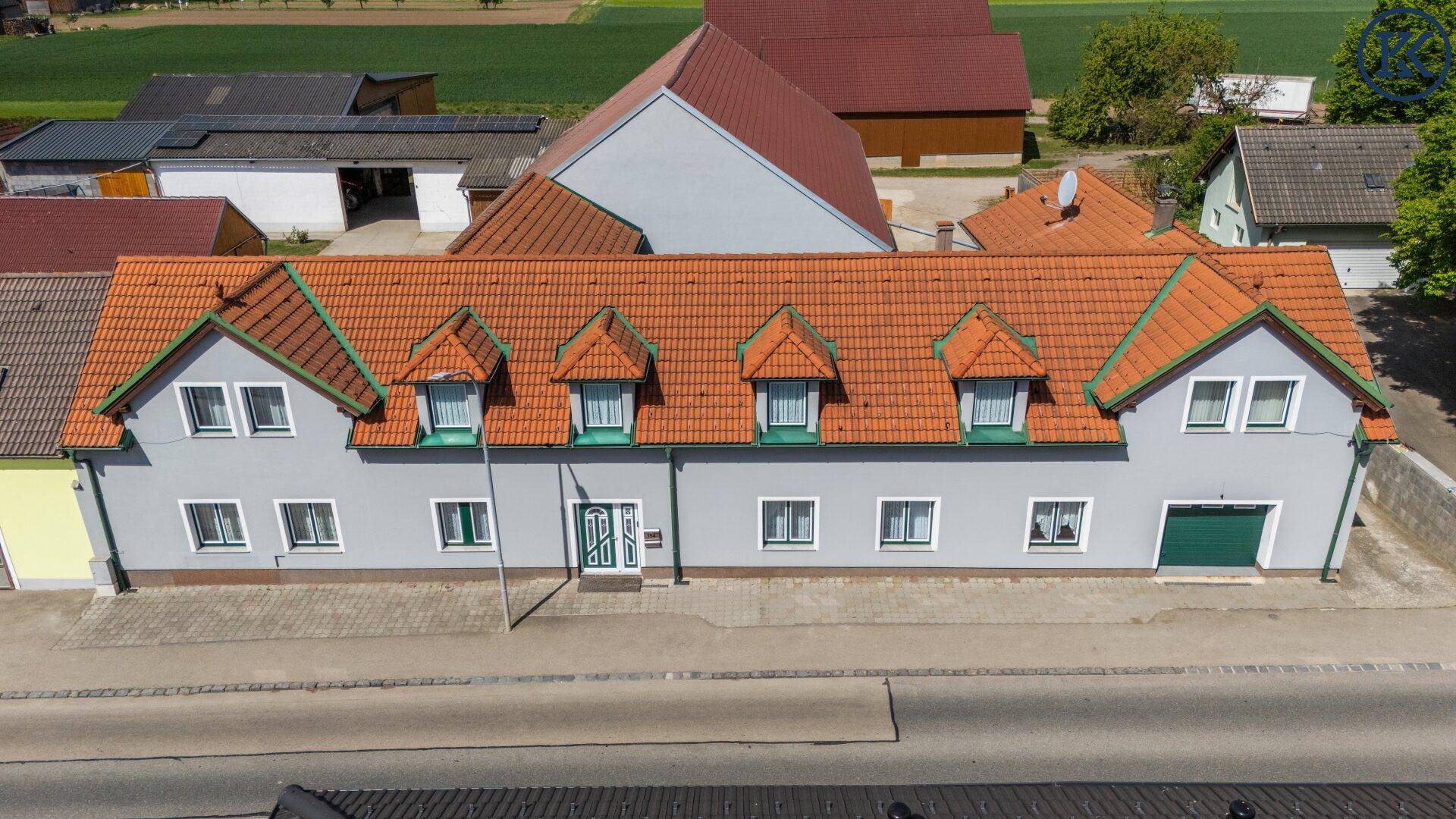 Luftaufnahme der Gebäudefassade mit rotem Ziegeldach, mehreren Gauben und angrenzender Straße.