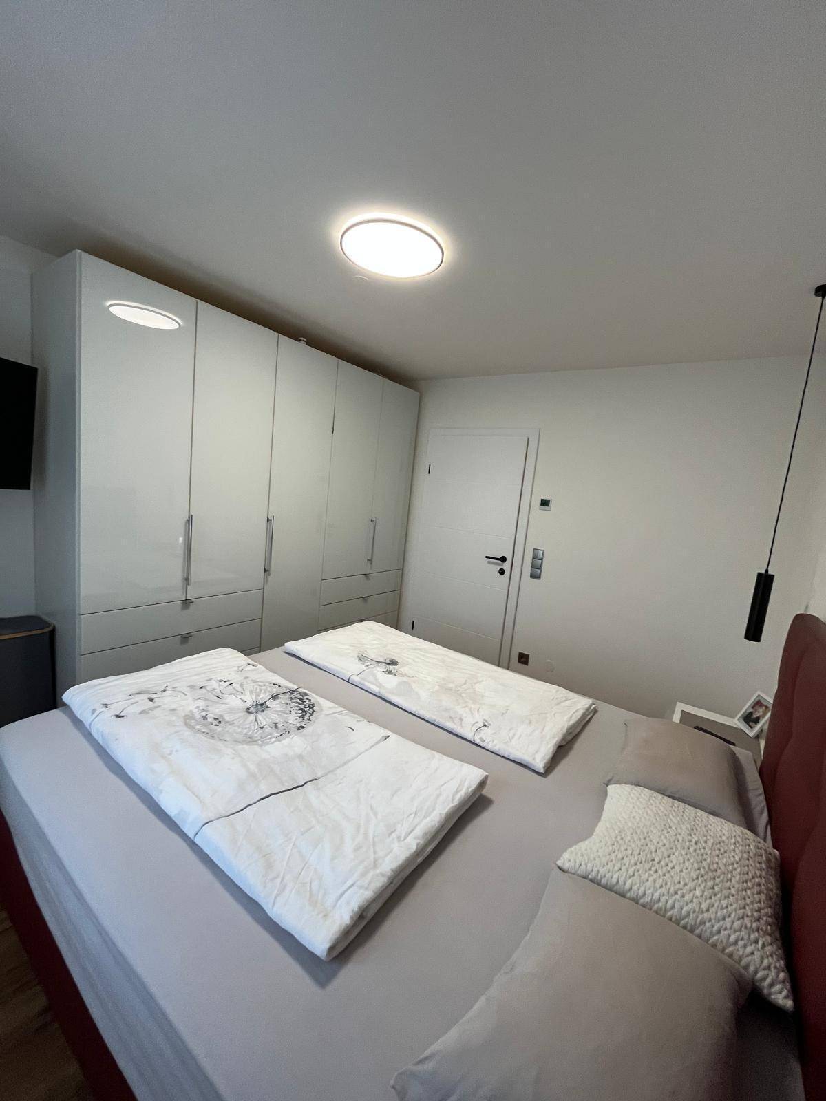 Schlafzimmer mit großem Bett, weißen Einbauschränken und moderner Deckenbeleuchtung.