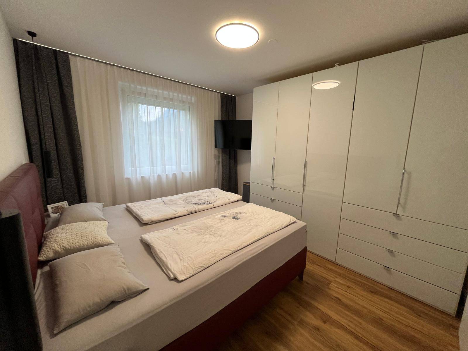 Schlafzimmer mit Doppelbett, großem weißen Kleiderschrank und Fenster mit Vorhängen.