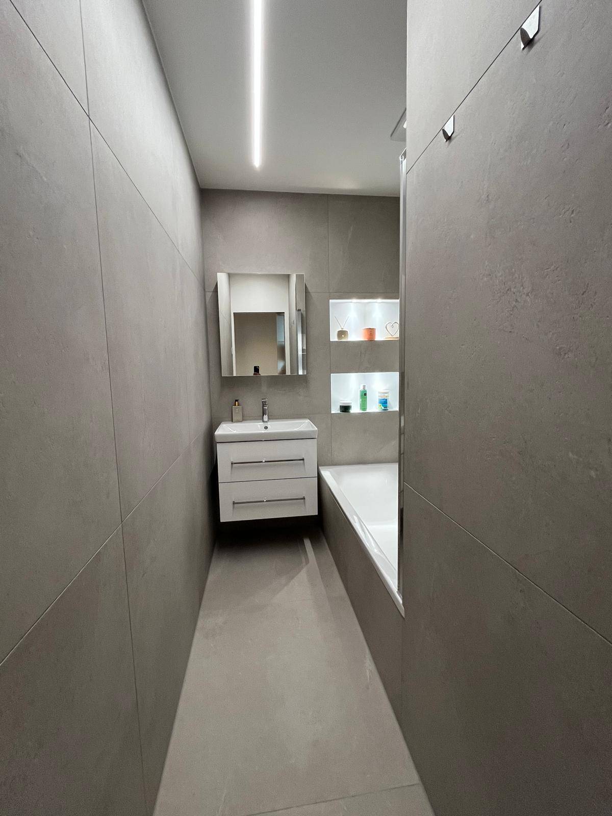 Modernes Badezimmer mit grauen Fliesen, Waschtisch und Badewanne mit Duschfunktion.