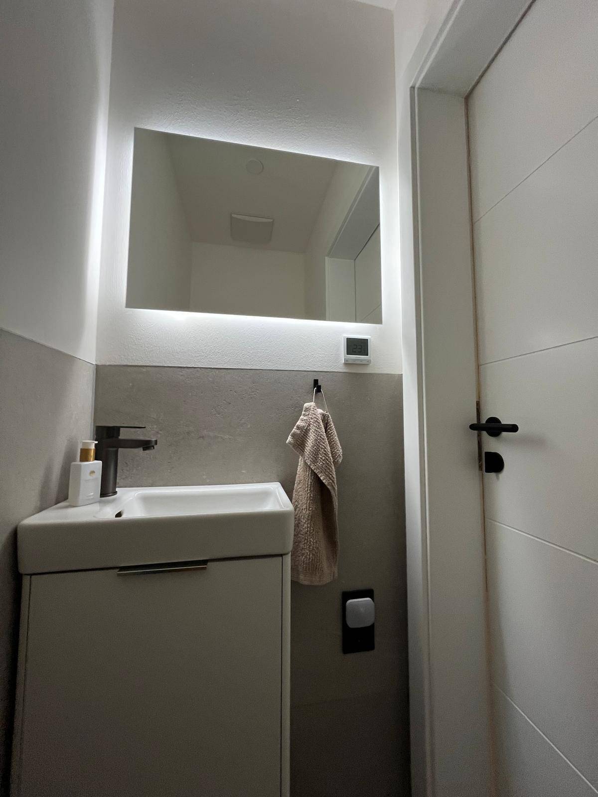 Gäste-WC mit modernem Waschbecken, beleuchtetem Spiegel und weißer Tür mit schwarzen Griffen.
