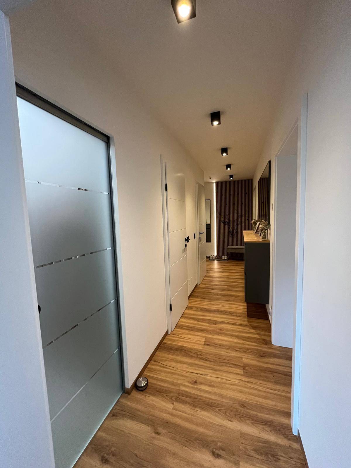 Langer Flur mit Laminatboden, Milchglastür und Blick auf die dekorative Holzlamellenwand.