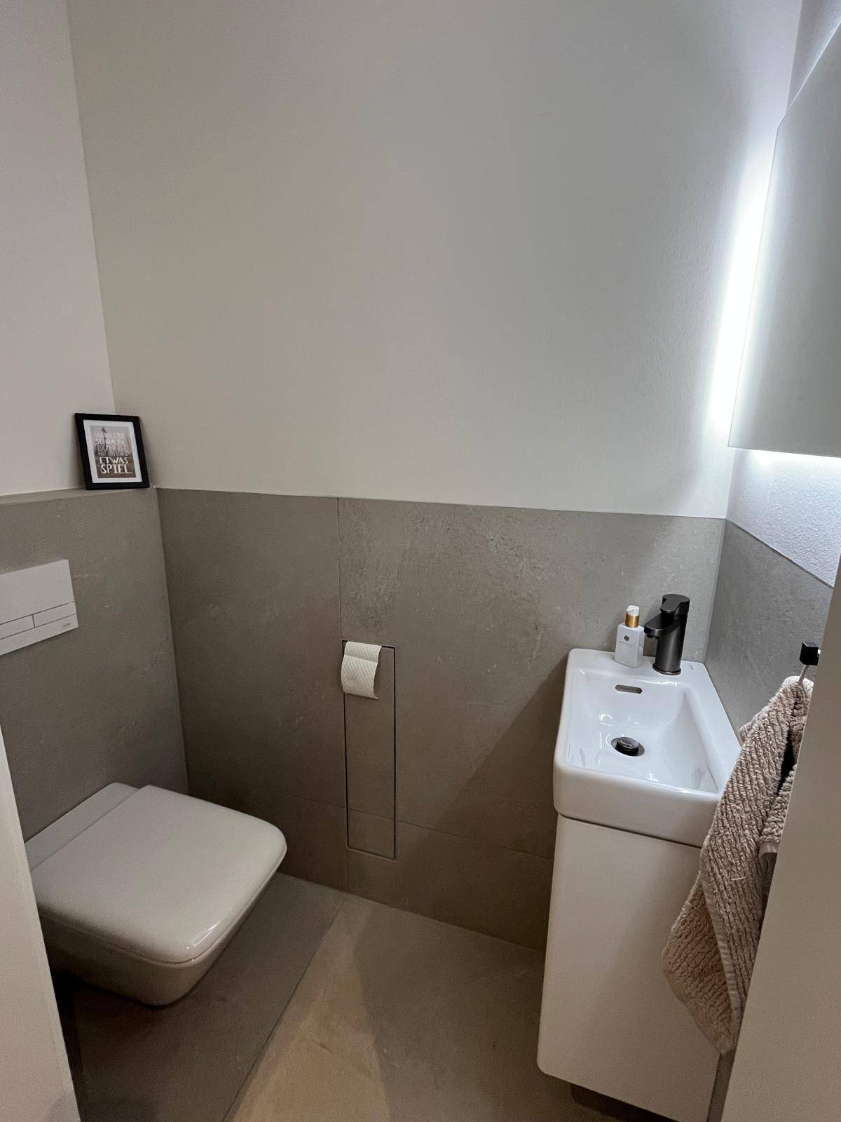 Modernes Gäste-WC mit wandhängender Toilette, kleinem Waschbecken und halbhohen grauen Fliesen.