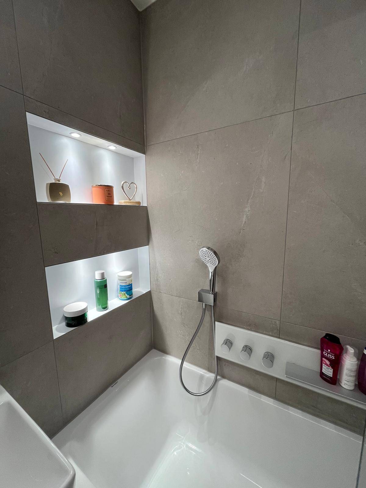 Modernes Badezimmer mit Badewanne, integrierten beleuchteten Nischen und grauen Fliesen an Wänden und Boden.