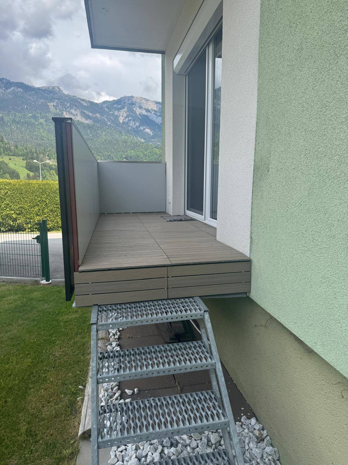 Balkon mit Holzboden und Metalltreppe zum Garten, umgeben von Bergen.