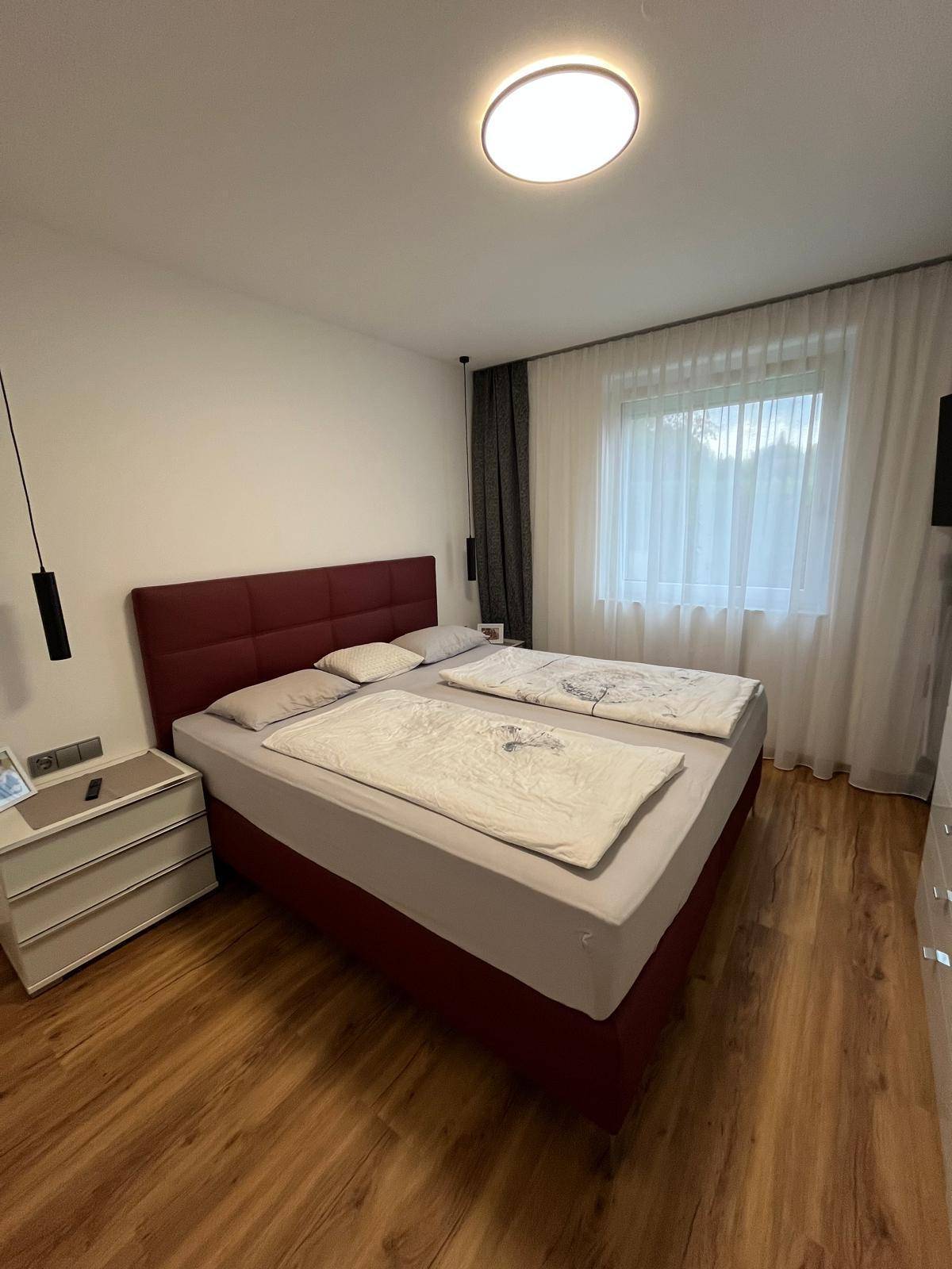 Gemütliches Schlafzimmer mit Doppelbett, Nachttisch und Fenster mit hellen Vorhängen.