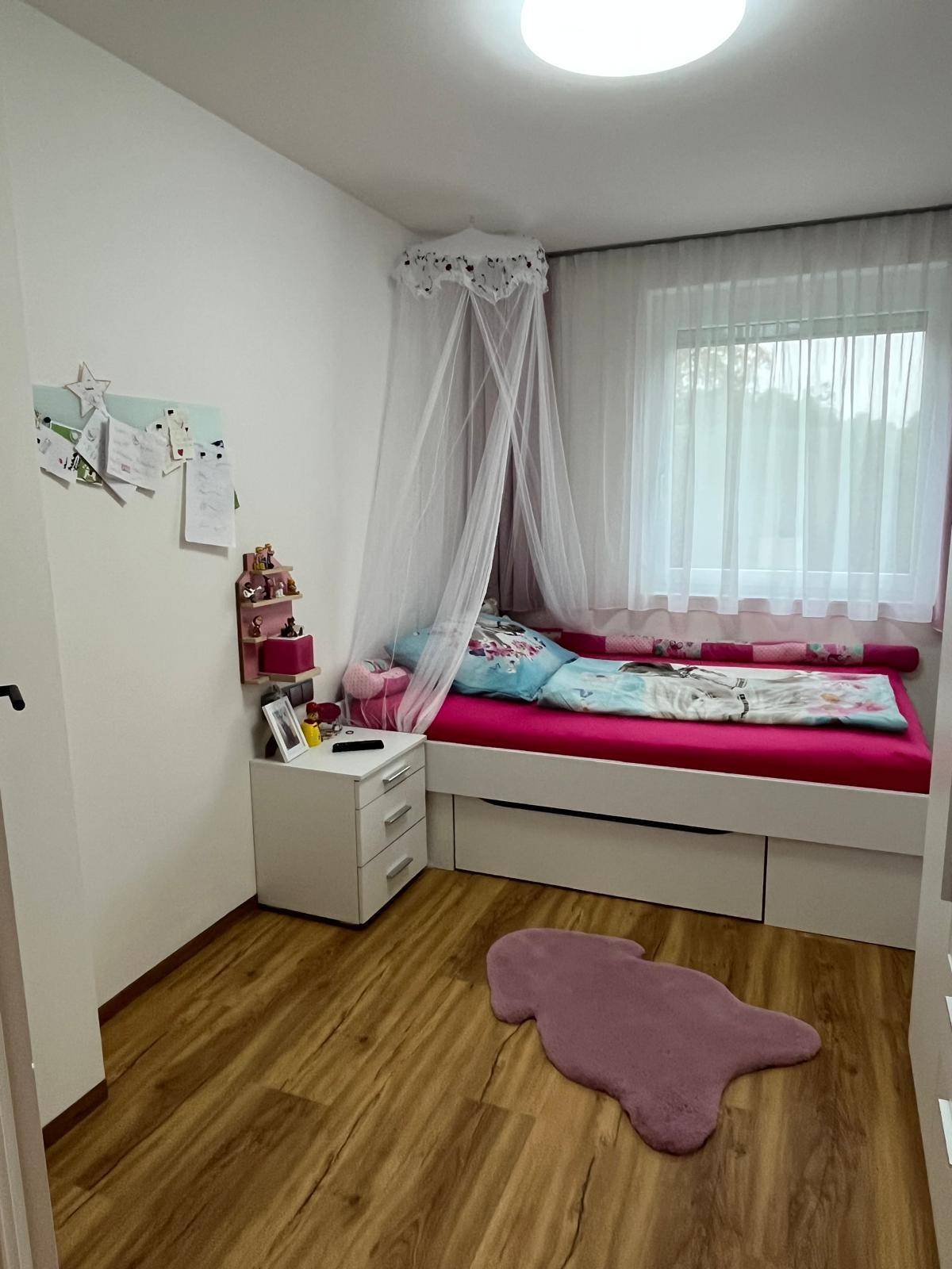 Helles Kinderzimmer mit Einzelbett, Himmelbett, rosa Bettwäsche und Fenster für Tageslicht.