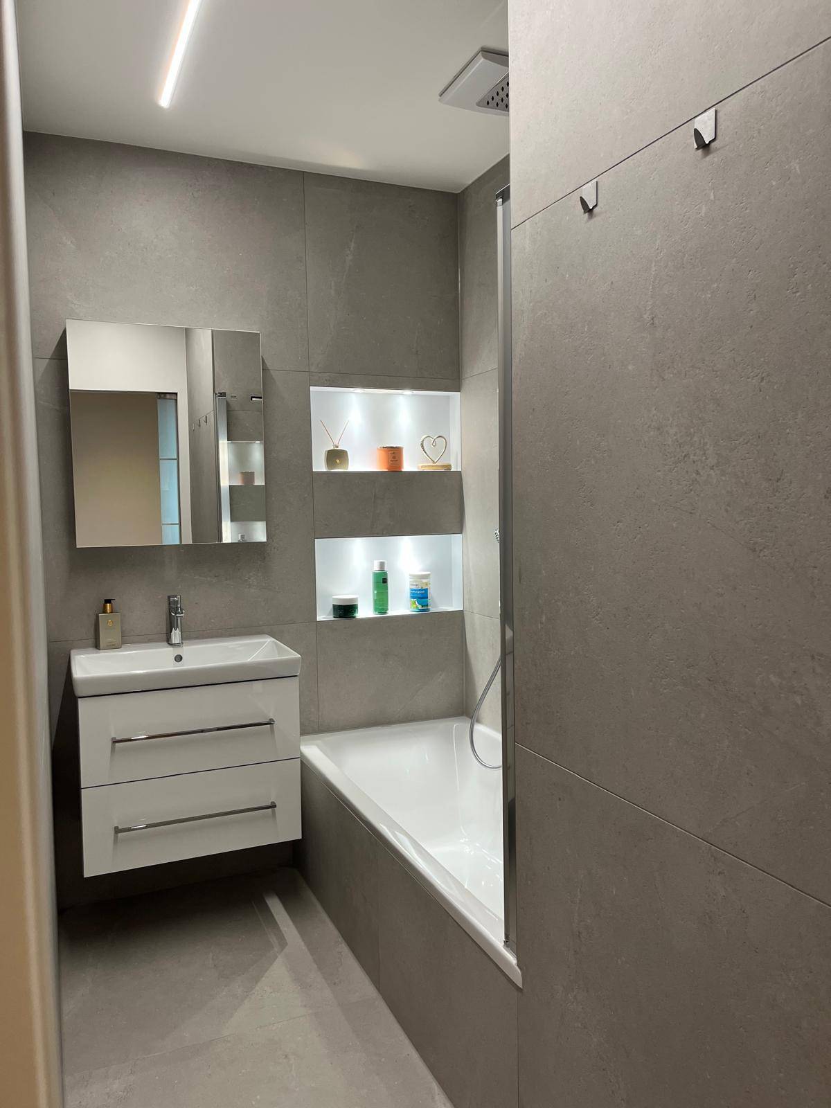 Modernes Badezimmer mit Badewanne, Waschtischunterschrank, Spiegelschrank und grauen Fliesen.