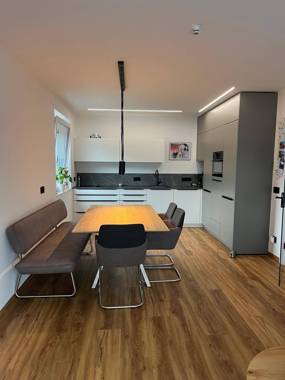 Moderner Essbereich mit Holztisch und Bank, offen zur Küche mit weißen und grauen Schränken.