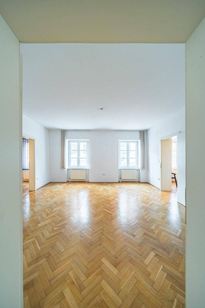 Heller Innenraum mit Fischgrätparkett und zwei großen Fenstern, gesehen durch einen Türrahmen.