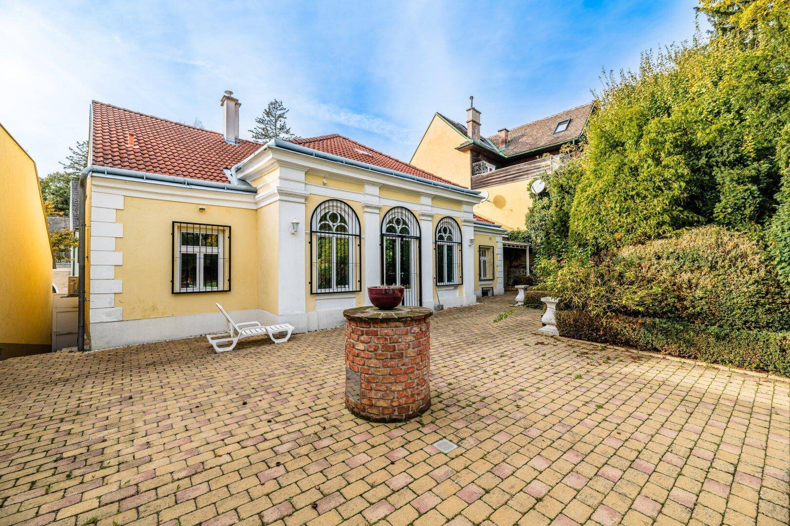 Historisches Gebäude mit gelber Fassade und gepflasterter Terrasse, umgeben von üppigem Grün.