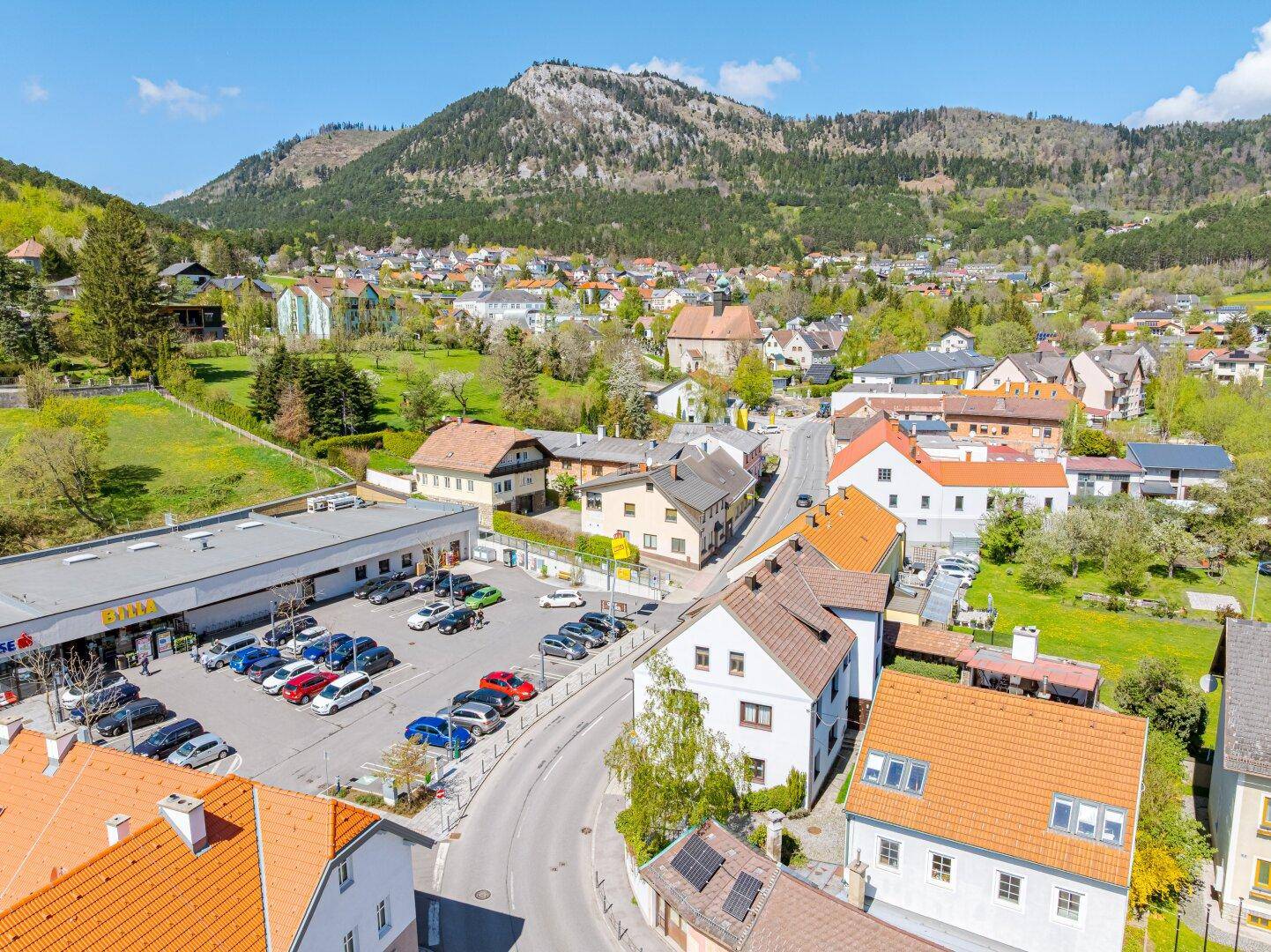 Panoramablick auf die Ortschaft mit Supermarkt, Wohngebäuden und majestätischer Bergkulisse.