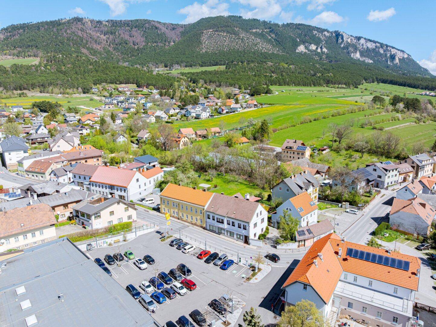 Weitläufige Ansicht der Siedlung mit Feldern, Bergen und einem Supermarkt im Vordergrund.