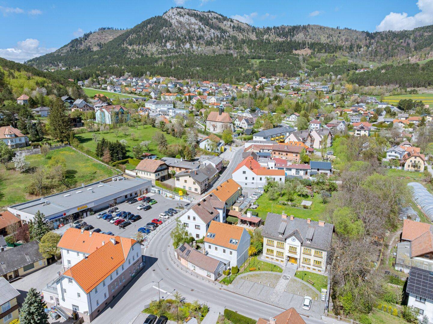 Luftaufnahme der Gemeinde mit Wohngebäuden, Supermarkt und beeindruckender Berglandschaft.