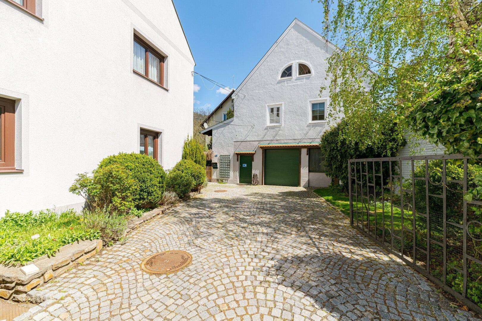 Gepflasterte Einfahrt zum hinteren Gebäude mit Garage und üppiger Bepflanzung.