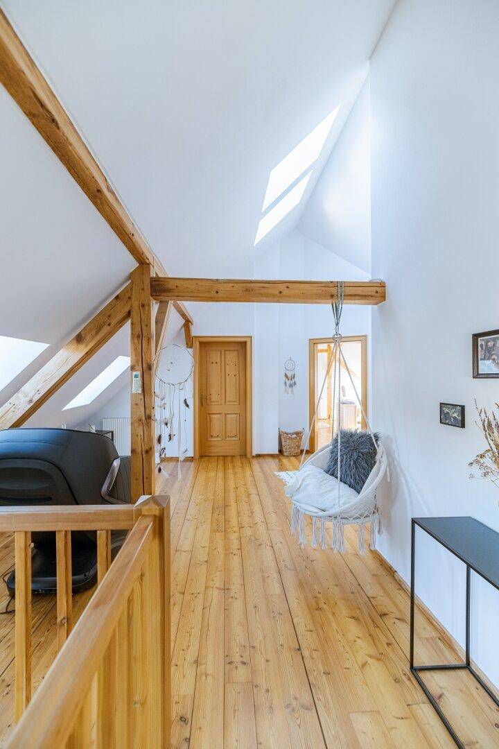 Offener Dachbodenbereich mit Holzbalken, Holzboden und Hängesessel unter dem Dachfenster.