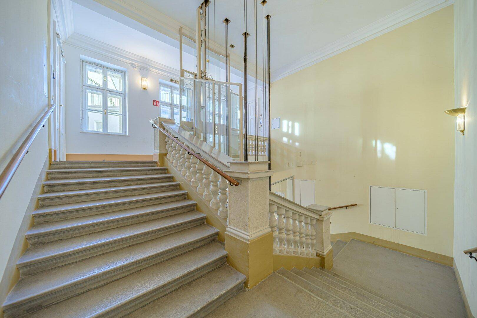 Klassisches Treppenhaus in einem Altbau mit sichtbarem Aufzugschacht, Balustrade und Fenstern.