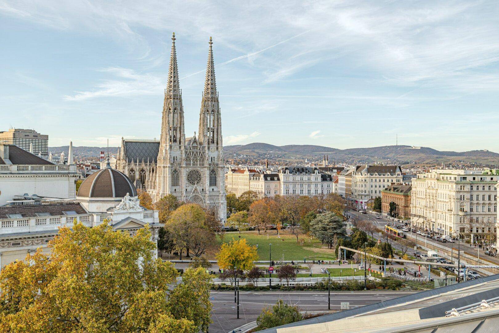 Weitläufiger Stadtblick mit der Votivkirche, einem grünen Park und umliegenden Gebäuden unter blauem Himmel.