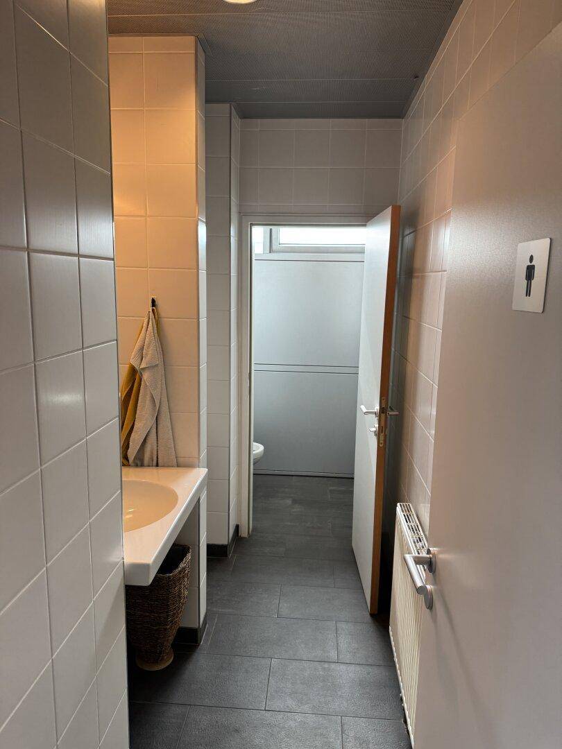 Schmaler Toilettenbereich mit hellen Fliesen an Wänden und Boden, der zu weiteren Kabinen führt.