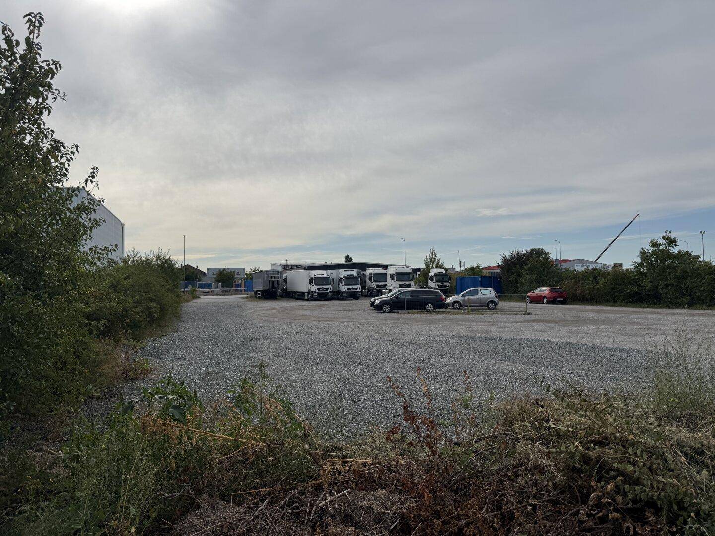 Weitläufiger Schotterparkplatz mit Lastwagen und Autos, umgeben von Grünflächen und Industriegebäuden.