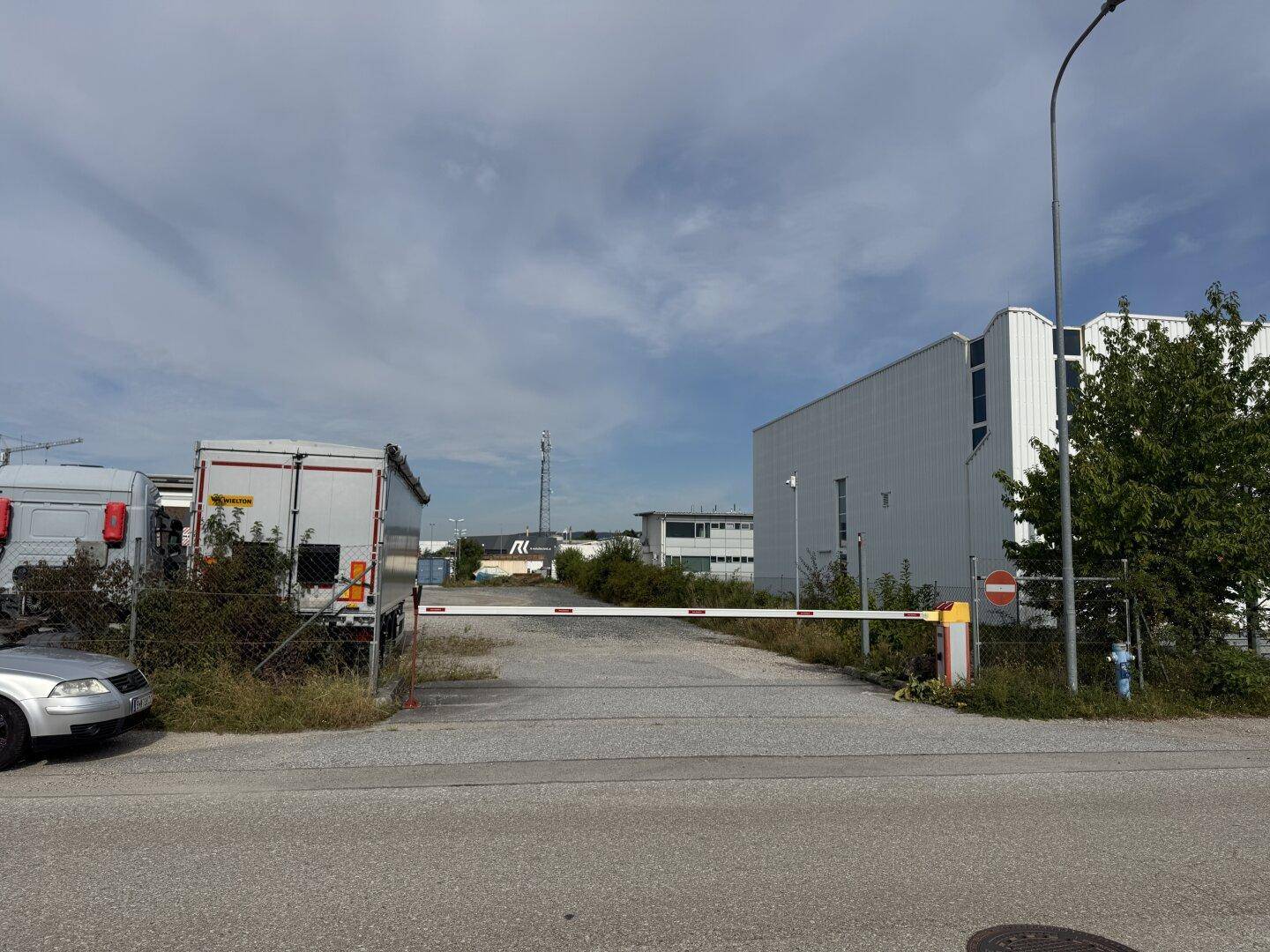 Eingangsbereich zu einem Industriegebiet mit einer Schranke, Schotterweg und Lagerhallen im Hintergrund.