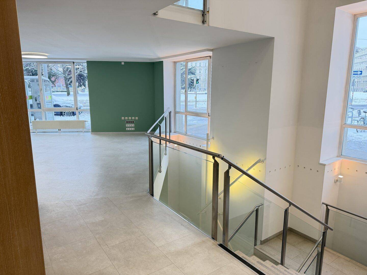 Heller Bürobereich mit großen Fenstern und Blick auf die Straße, angrenzend an einen Treppenaufgang.