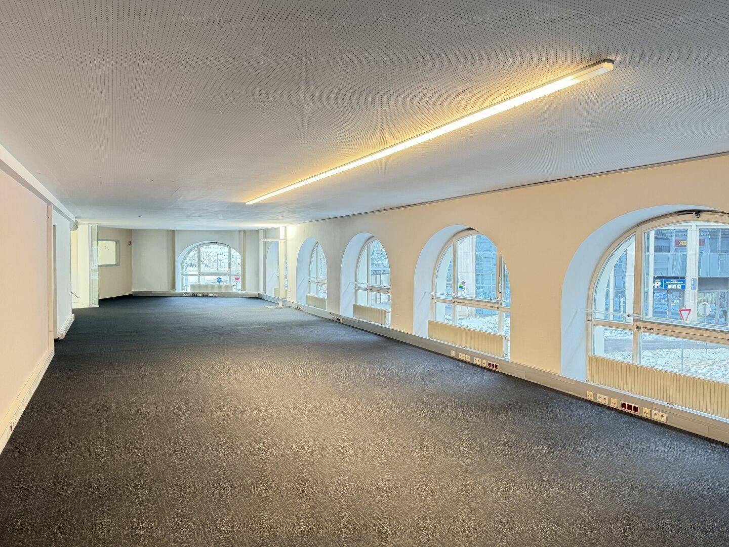 Großer Büroraum mit Teppichboden und charakteristischen Rundbogenfenstern, die zur Straße zeigen.