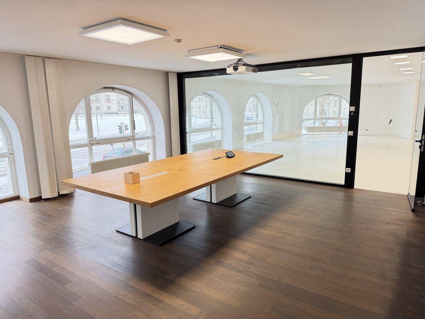 Moderner Konferenzraum mit großem Holztisch, dunklem Holzboden und Projektor, durch Glaswand abgetrennt.