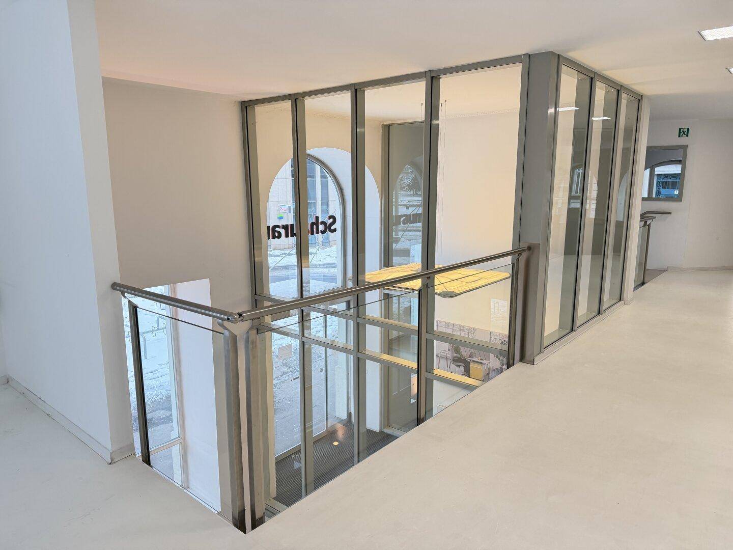 Flur mit Glasgeländer und Blick auf einen Aufzug oder eine weitere Etage, hell und modern gestaltet.