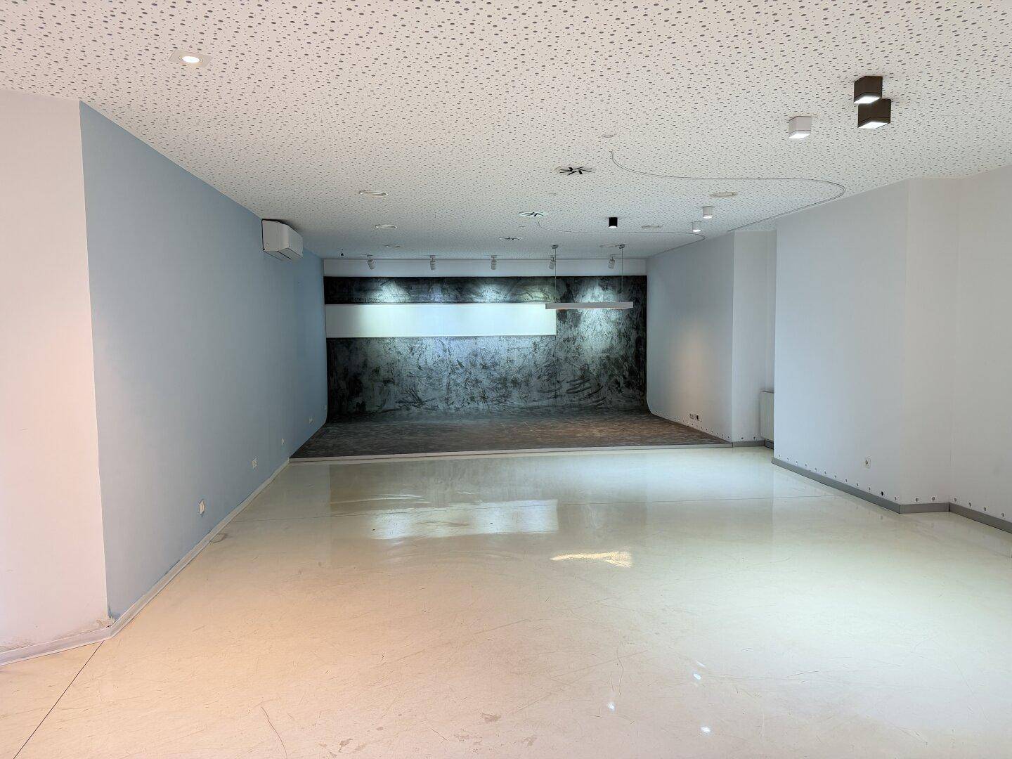Großer, leerer Büroraum mit hellem Boden, Akustikdecke und einer strukturierten Wand im Hintergrund.