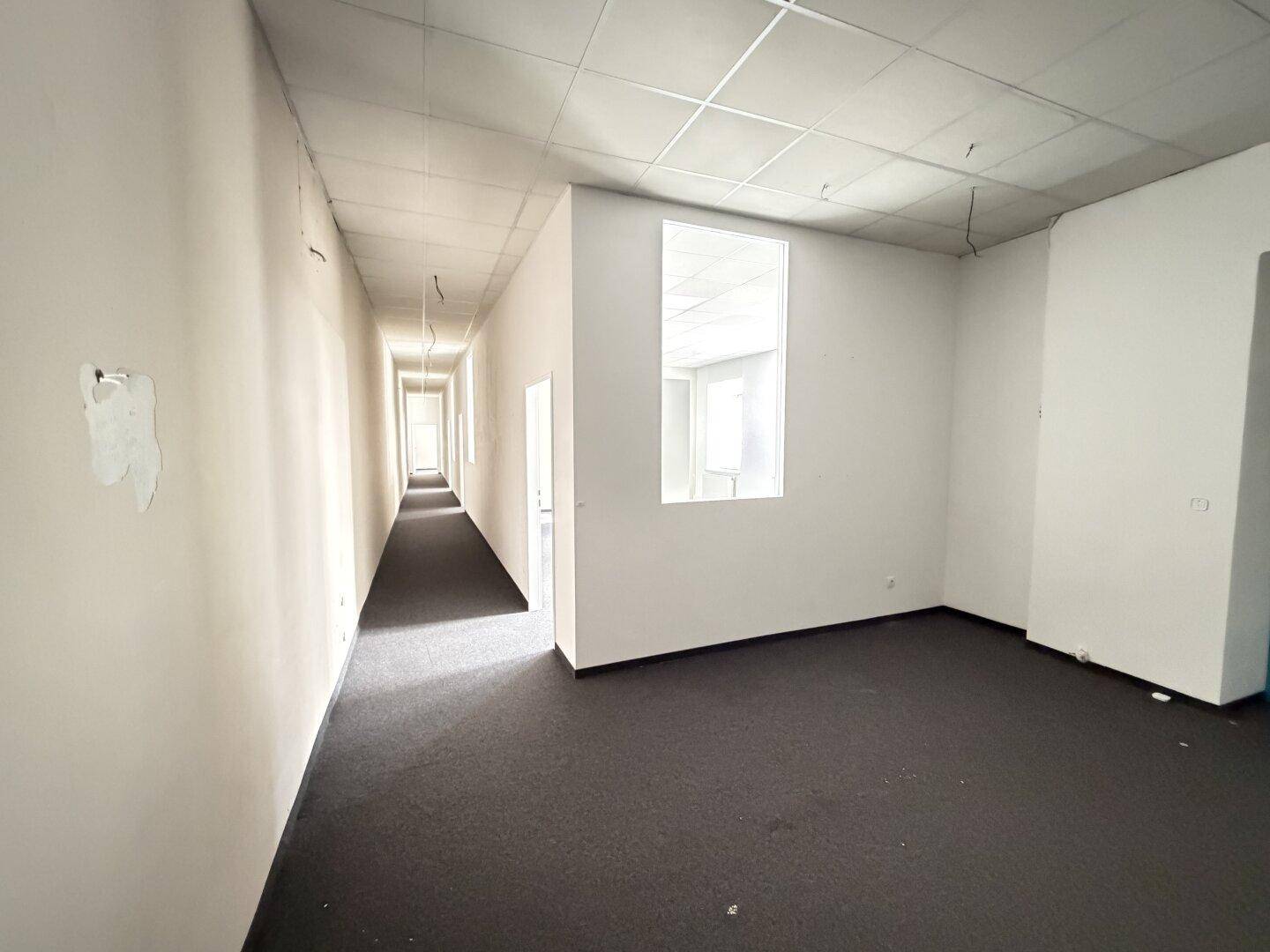 Leerer Büroraum mit Teppichboden und einem Durchgang zu einem langen Flur, hell beleuchtet.